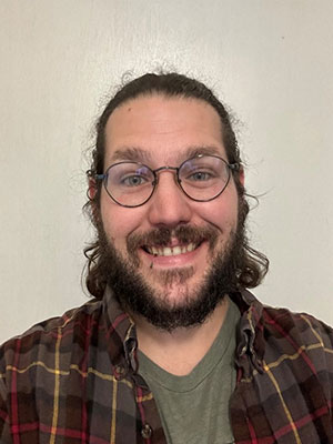 White Man longer brown hair and brown beard. Glasses red and brown plaid shirt. Smiling in front of plain wall