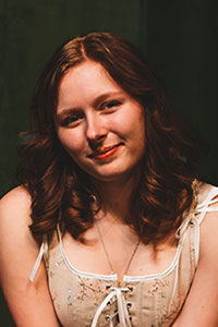 woman with white floral bluse sitting in front of a dark green curtain under professional lighting smiling