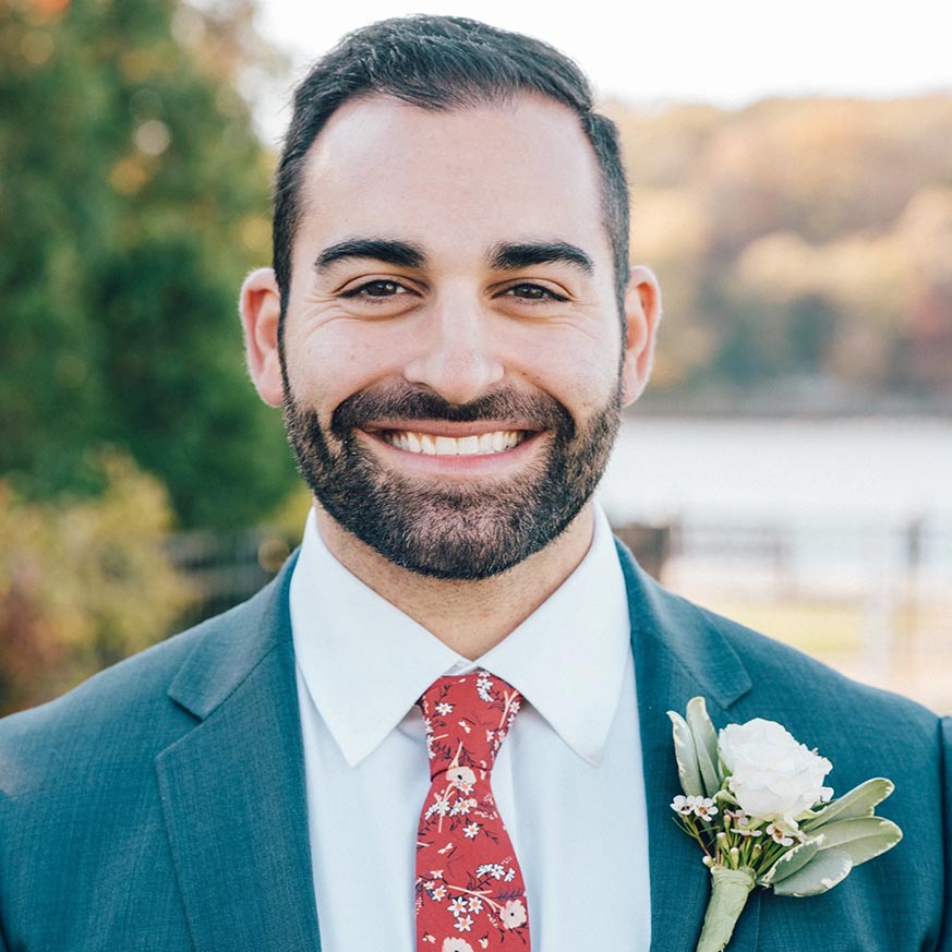 man with dark hair and beard wearing a blue suit and floral tie