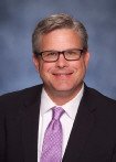 White man wearing glasses, a black suit, and purple tie