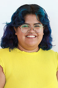 Woman with olive skin and shoulder length curly black hair with blue and purple pieces wearing a yellow shirt and glasses