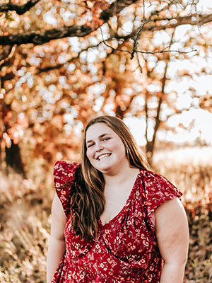 White woman with long brown hair wearing a floral read dress posing outside