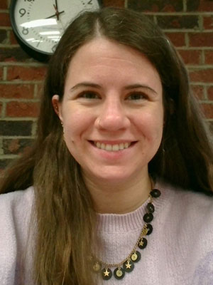 White woman with long brown hair smiling wearing a pastel sweater and gold and black necklace sitting in from of an interior brick wall