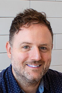 Man with a grey beard and brown hair and blue eyes wearing a dark blue collared shirt